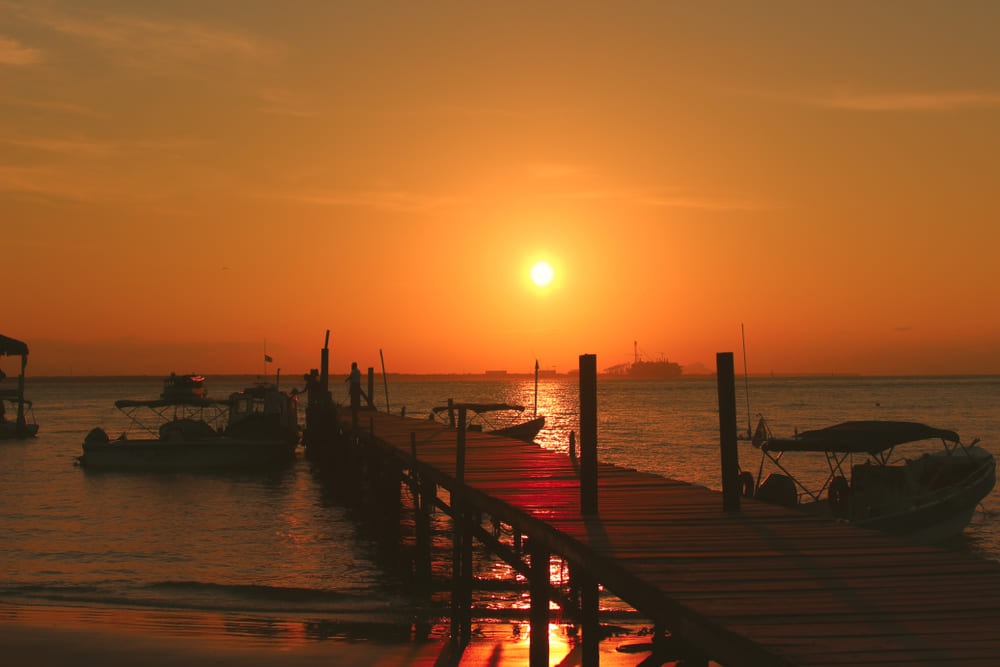 Foto de um lindo pôr do sol na Ilha do Mel, no Paraná, onde é possível ver o mar, os barcos e um píer.