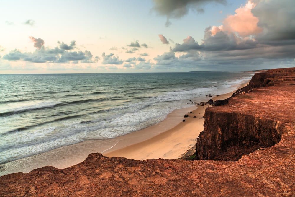 Foto da praia de Pipa, no Rio Grande do Norte, com o morro ao lado e a praia ao fundo. O mar está agitado e já está entardecendo.