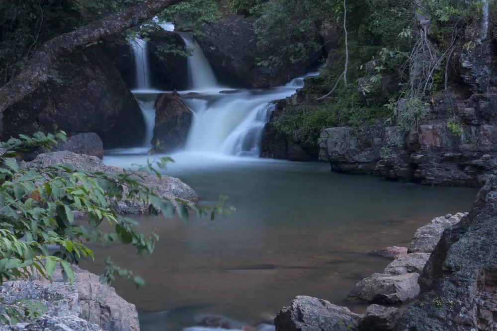 Foto de uma das infinitas cachoeiras que ficam na região de Pirenópolis, Goiás. A cachoeira é pequena, mas com uma forte queda d'água. O dia parece estar entardecendo e a água mescla de verde para marrom.