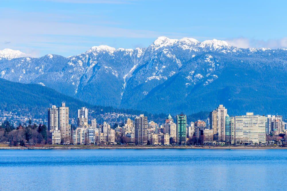 Foto de uma das regiões mais famosas de Vancouver no Canadá. É possível ver muitos prédios na orla do mar e as rochas e montanhas enormes e com seus picos nevados ao fundo.