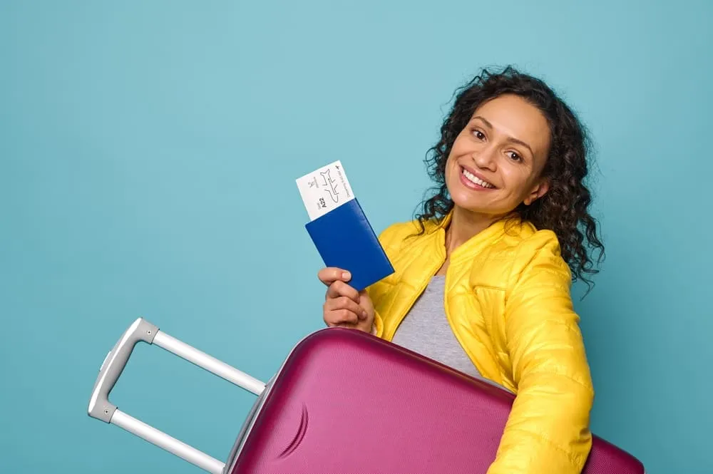 Foto de uma mulher sorrindo com uma mala de viagem e seus documentos pessoais nas mãos.