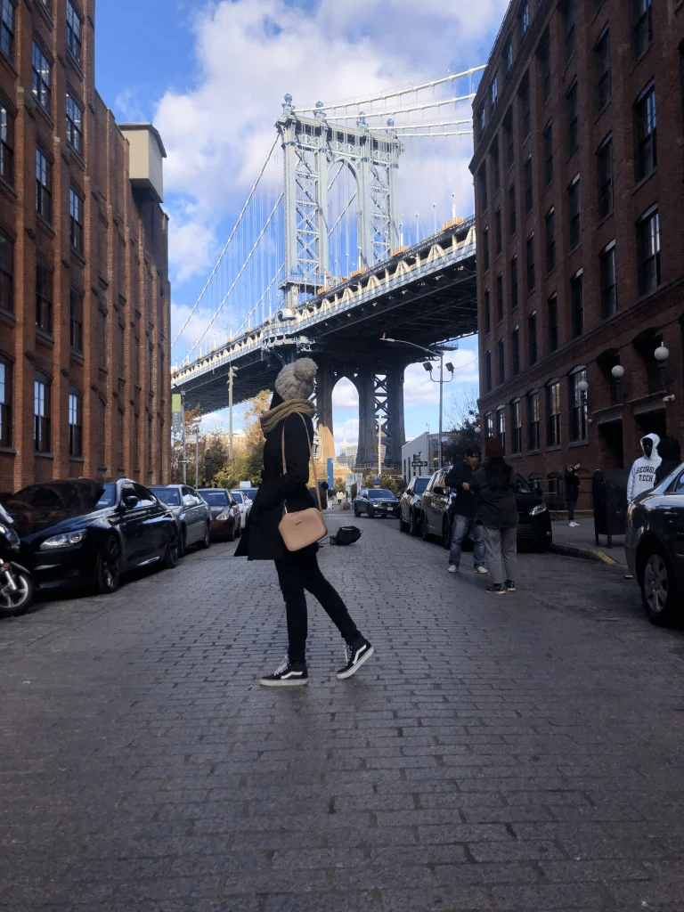 Foto da ponte do Brooklyn, onde é possível ver uma mulher em frente e a ponte ao fundo. Há pessoas ao redor e carros estacionados. O céu está azul.