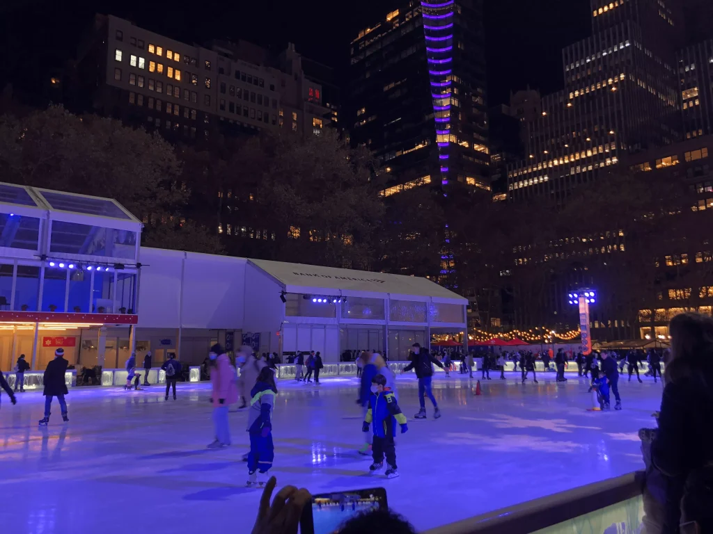 Foto da pista de gelo presente no Bryant Park em New York. Há pessoas patinando no gelo e os prédios ao redor estão com as luzes acesas já que a foto é noturna.