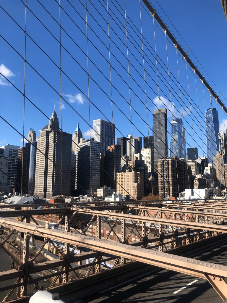 Foto da ponte do Brooklyn, onde é possível veros prédios de Manhattan, New York, ao fundo. O céu está azul.
