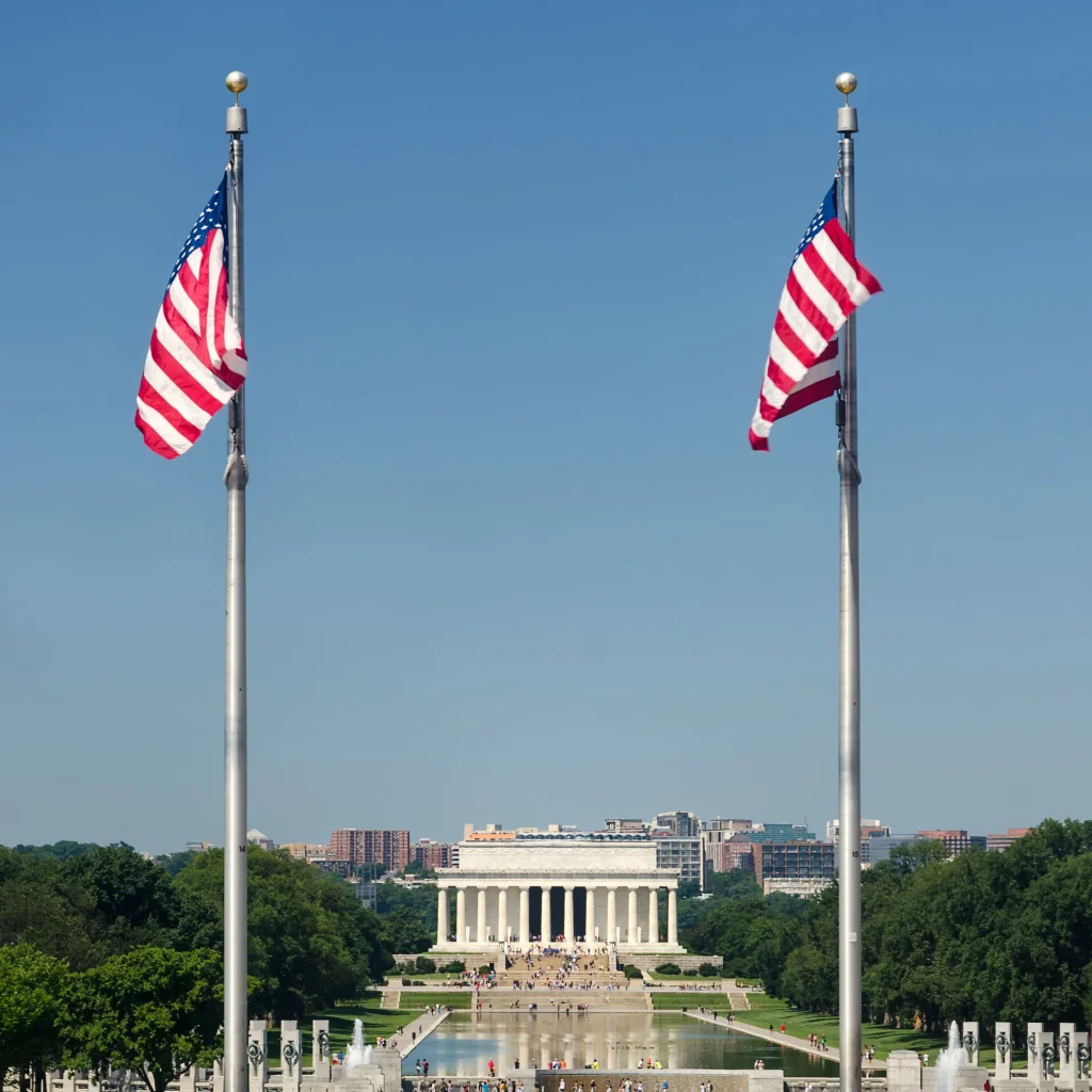 Foto da Casa Branca na cidade de Washington D.C. com as bandeias dos Estados Unidos. O céu está azul e há pessoas transitando pelo jardim do local.