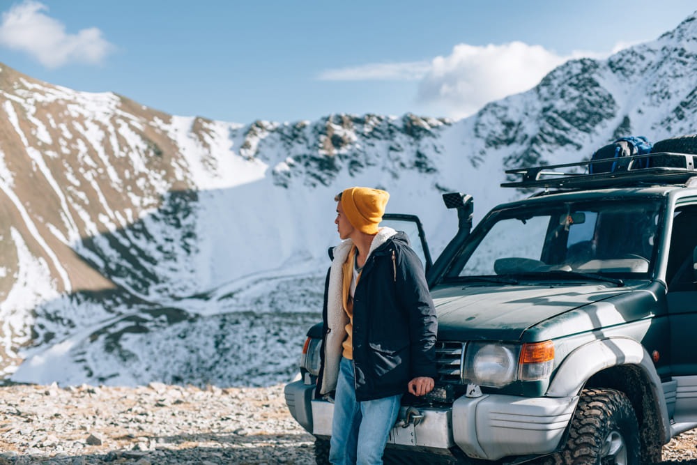 Foto de um homem bem agasalhado em frente ao seu carro alugado. Ao fundo, é possível ver os picos montanhosos repletos de neve.