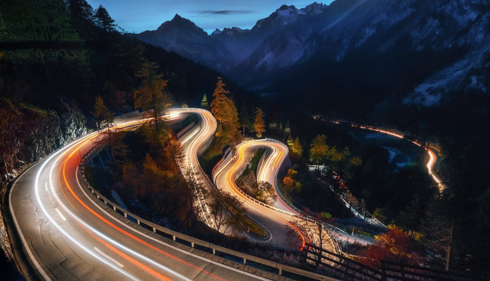 Foto de uma estrada super sinuosa, repleta de curvas bem fechadas. Está de noite e é possível ver as luzes dos carros em alta velocidade passando.