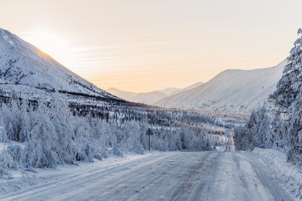 Foto da estrada de Kolyma, onde o percurso inteiro é repleto de neve. As árvores da estrada estão repletas de neve também. O sol está se pondo.