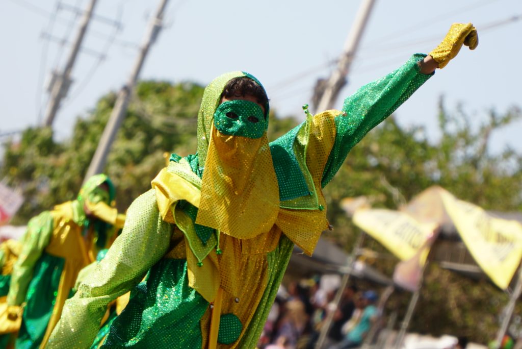 Figura del Monocuco en el Carnaval de Barranquilla, vistiendo un traje brillante en verde y amarillo con máscara y capucha, levantando un brazo en medio del desfile