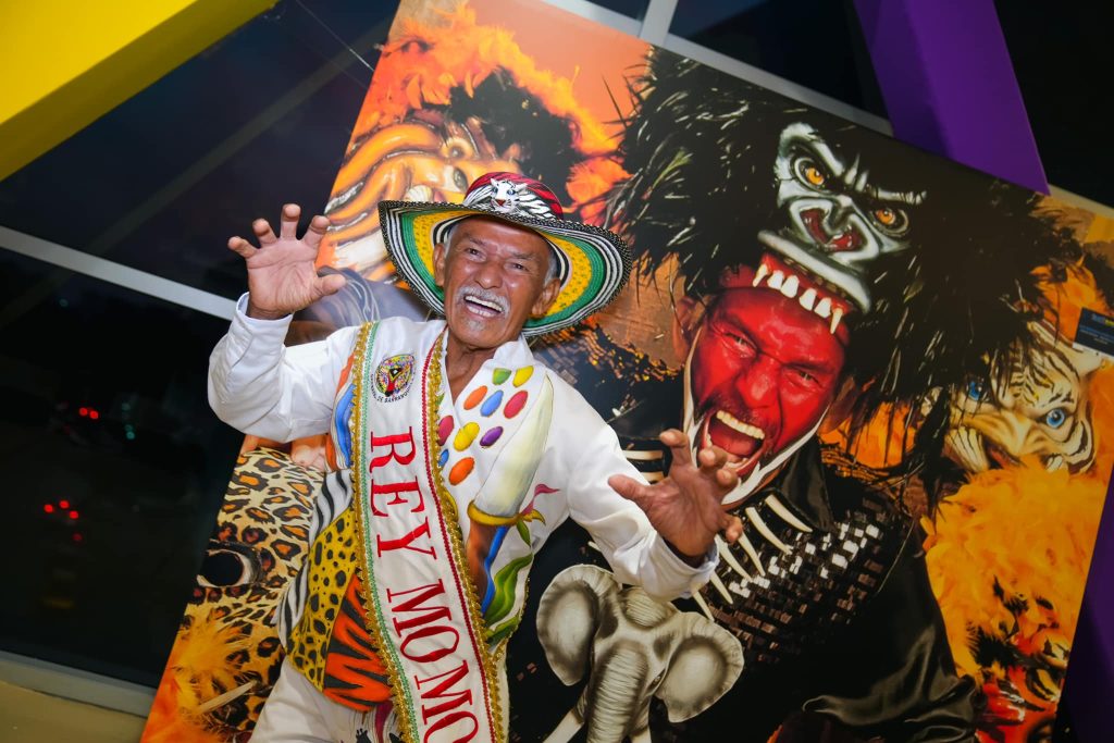 Rey Momo en el Carnaval de Barranquilla posando con entusiasmo, vistiendo un sombrero colorido y un traje tradicional, frente a un mural vibrante con máscaras tribales y figuras carnavalescas.