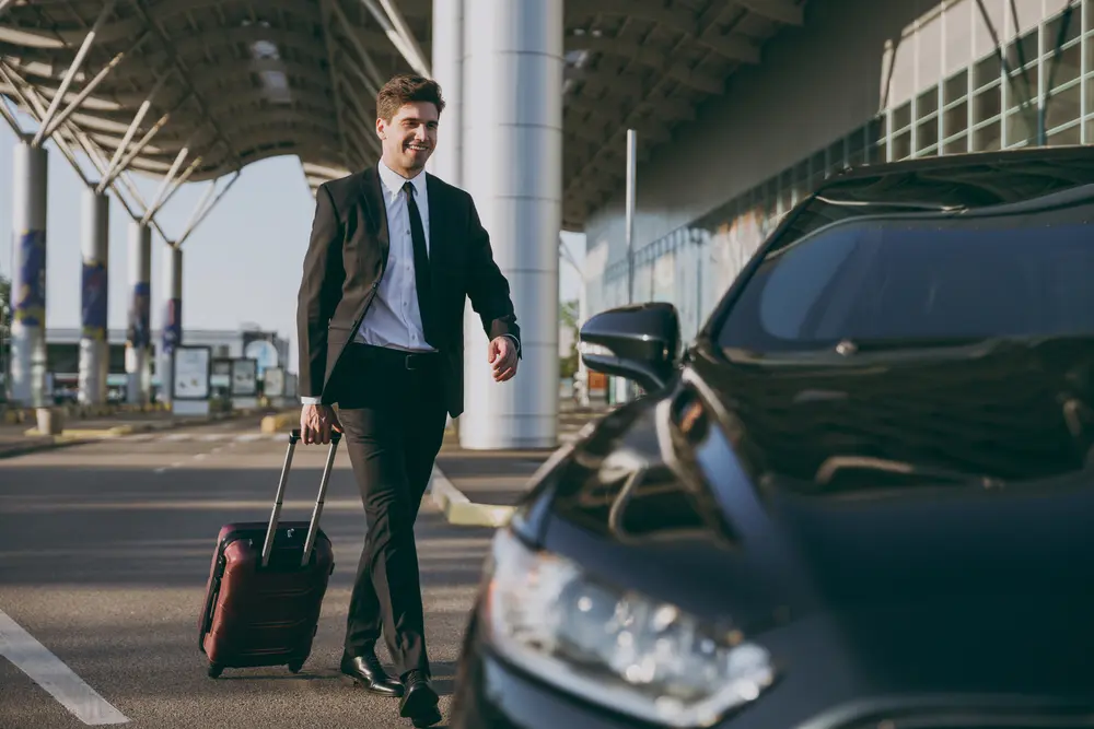 Foto de hombre en traje saliendo del aeropuerto con su maleta en dirección a su auto rentado para hacer turismo de negocios.