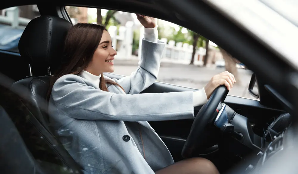 Mujer vistiendo blazer gris y sonriendo mientras maneja el auto que rentó para hacer turismo de negocios.