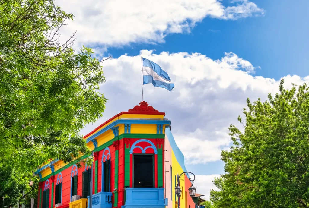 Foto do ponto turístico famoso da região de Buenos Aires, na Argentina, o El Caminito, com suas cores super chamativas. Ao topo, vemos a bandeira do país. O dia está claro.