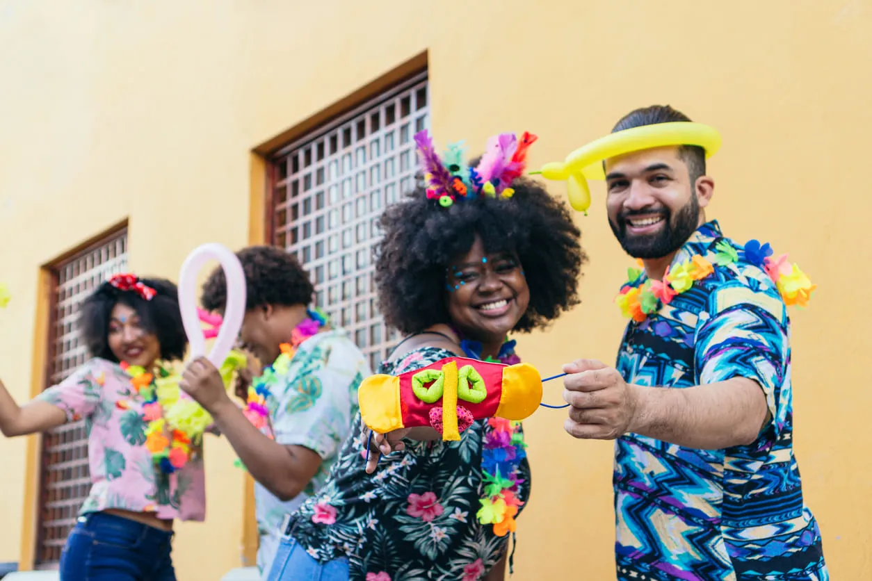 Foto de quatro pessoas comemorando o Carnaval. As roupas são coloridas e eles carregam elementos da festa: bexiga colorida, máscaras coloridas e acessórios para o cabelo. Todos sorriem para foto.