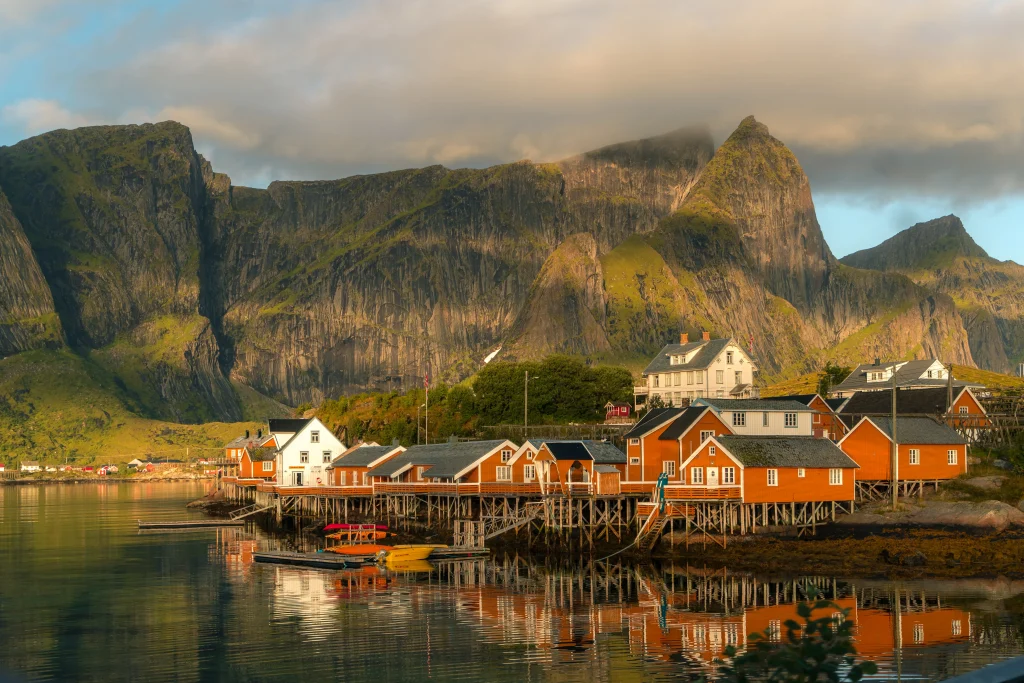 Foto de uma pequena vila na Escandinávia, onde é possível ver várias casas na cor laranja, todas padronizadas. Um lago fica em frente e montanhas enormes ao fundo.