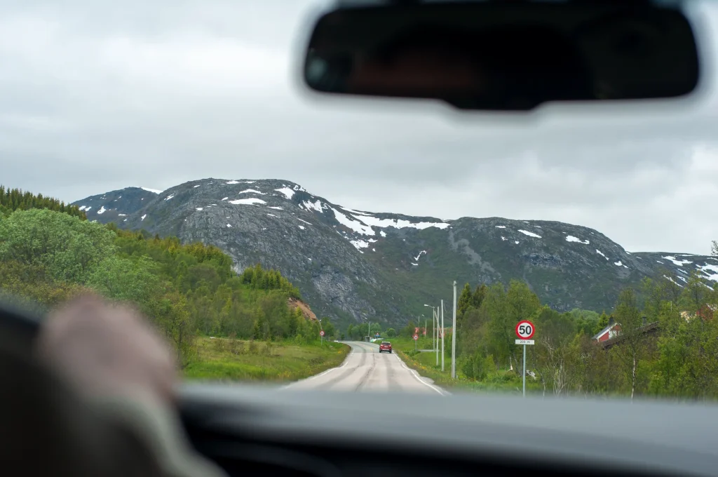 Foto de dentro do carro, onde é possível ver a estrada em frente e os picos nevados ao fundo. A estrada fica na Noruega.