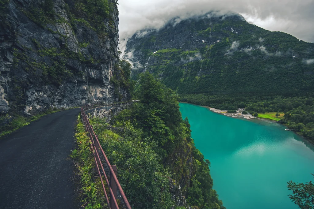 Foto incrível de um lago de cor azul bem forte na Noruega. É possível ver a estrada ao lado, a vegetação entre o lago e a estrada, e as montanhas enormes ao fundo.