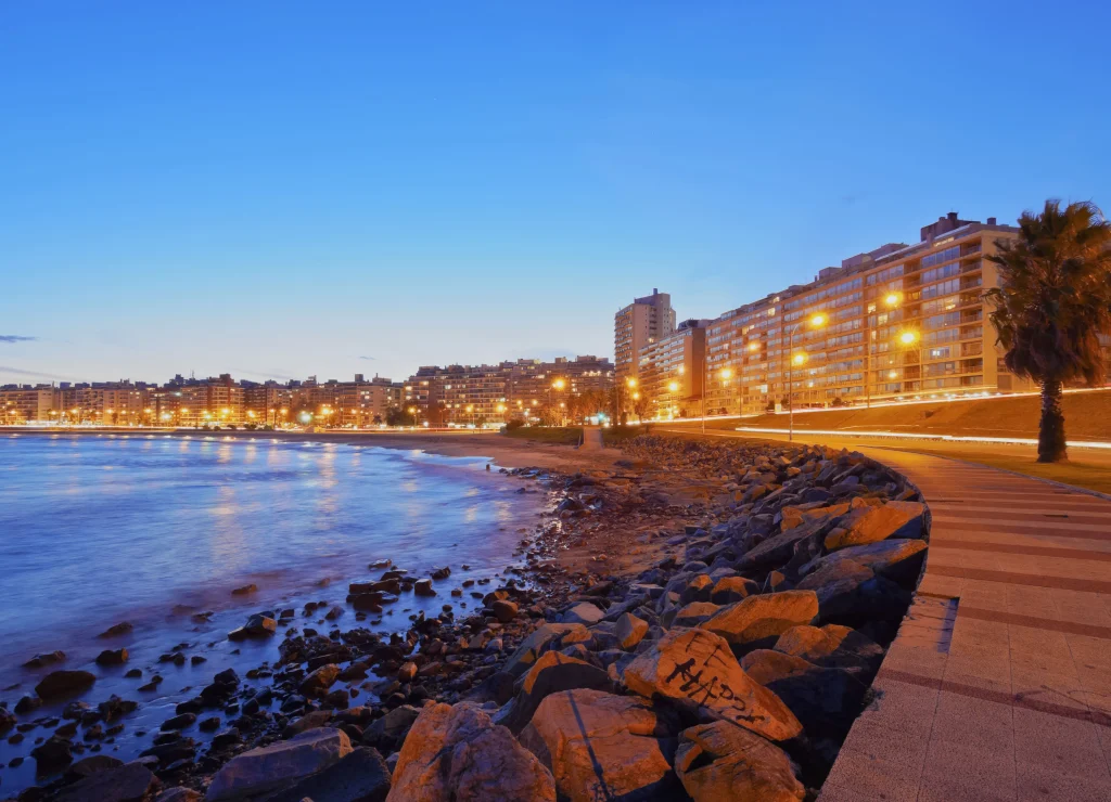 Foto da orla da praia na cidade de Montevidéu, Uruguai. A noite está começando a aparecer, o céu está azul e é possível ver as luzes de todos os prédios ao fundo.