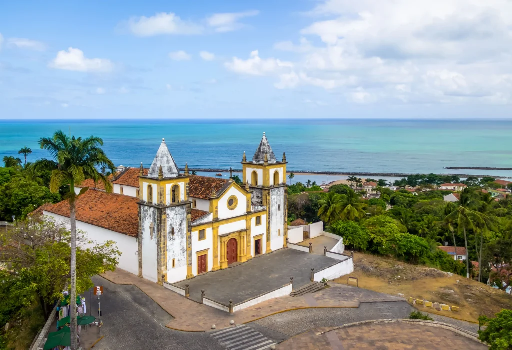 Foto aérea de Olinda, Pernambuco. É possível ver a Igreja de Nossa Senhora do Carmo com o mar vasto e azul ao fundo. A Igreja é rústica e pintada de branco e amarelo.