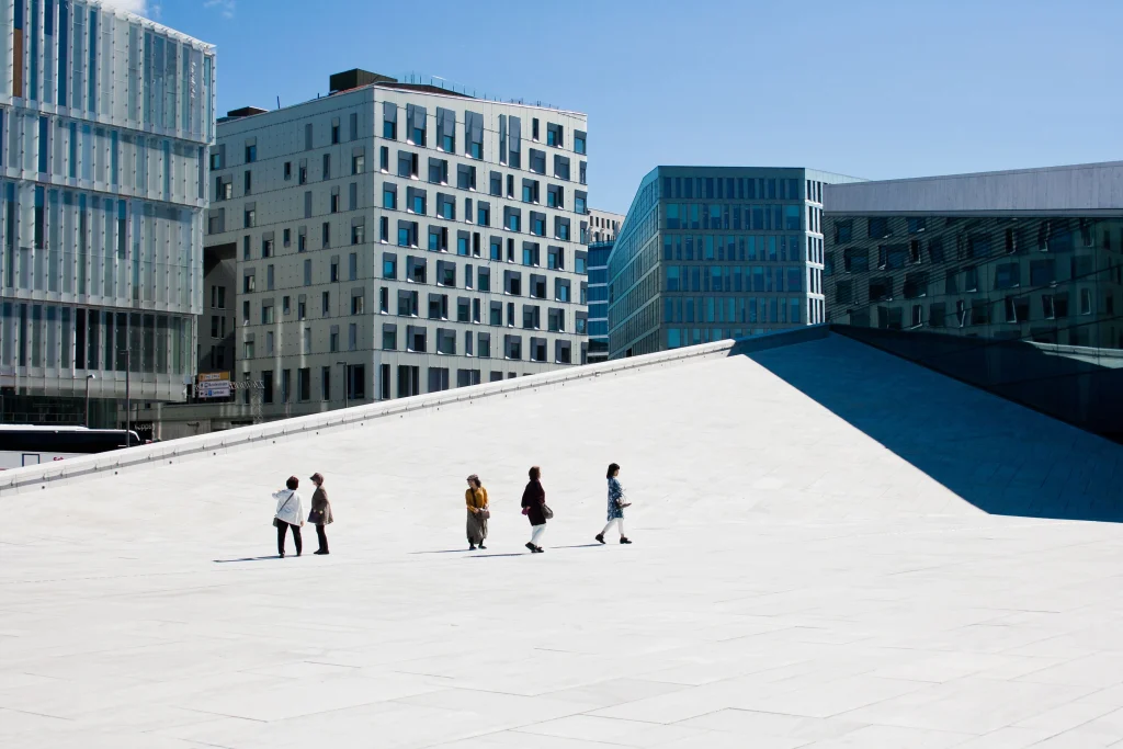 Foto de pessoas andando por uma dos pontos turísticos da cidade de Oslo, na Noruega. Ao fundo, é possível ver prédios com arquitetura futurística e moderna. O céu está super azul.