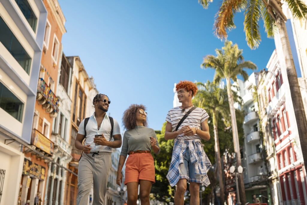 Foto de três amigos passeando pelas ruas de uma cidade. O céu está super azul e os prédios são coloridos.