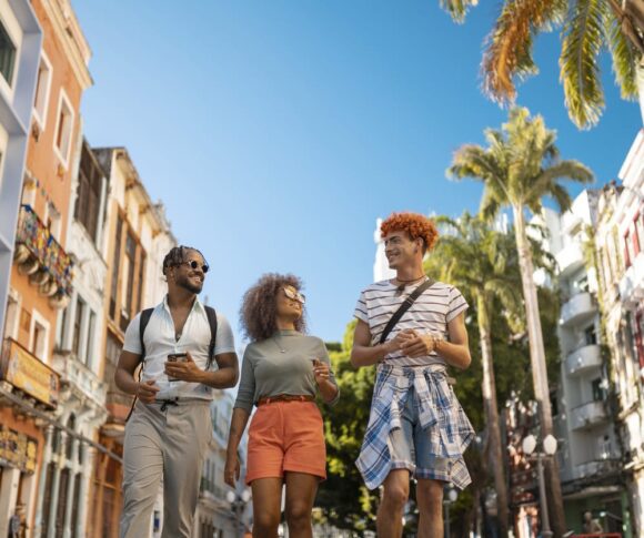 Foto de três amigos passeando pelas ruas de uma cidade. O céu está super azul e os prédios são coloridos.