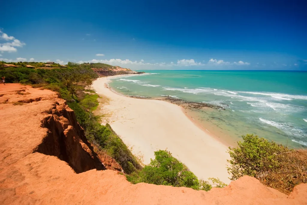 Foto da Praia de Pipa, no Rio Grande do Norte. A muralha de terra vermelha fica de um lado e o mar azul do outro, sendo que a separação se dá pela vasta faixa de areia. Ainda há algumas vegetações na praia e a areia está vazia.