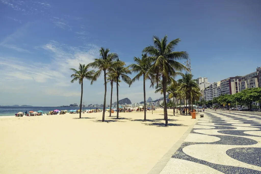 Foto da orla de Copacabana, no Rio de Janeiro. A foto mostra a vasta faixa de areia e as árvores da região. O céu está azul e tem diversos guarda-sóis montados na areia.