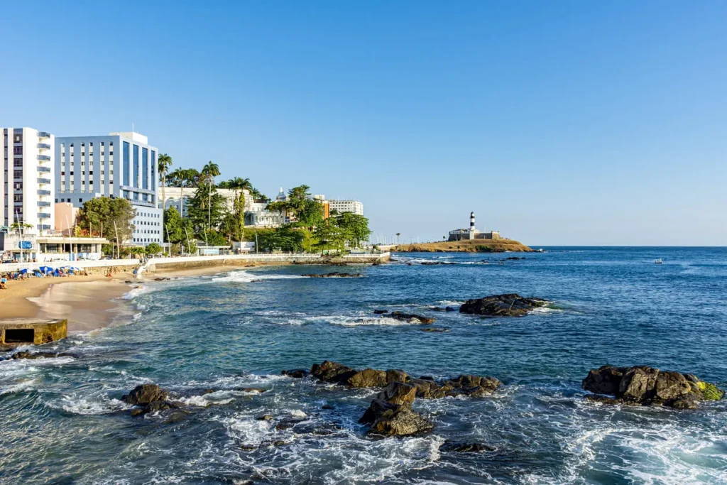 Foto da praia do Forte, em Salvador, na Bahia. Além do mar azul e suas pedras, é possível ver o Farol da Barra ao fundo.