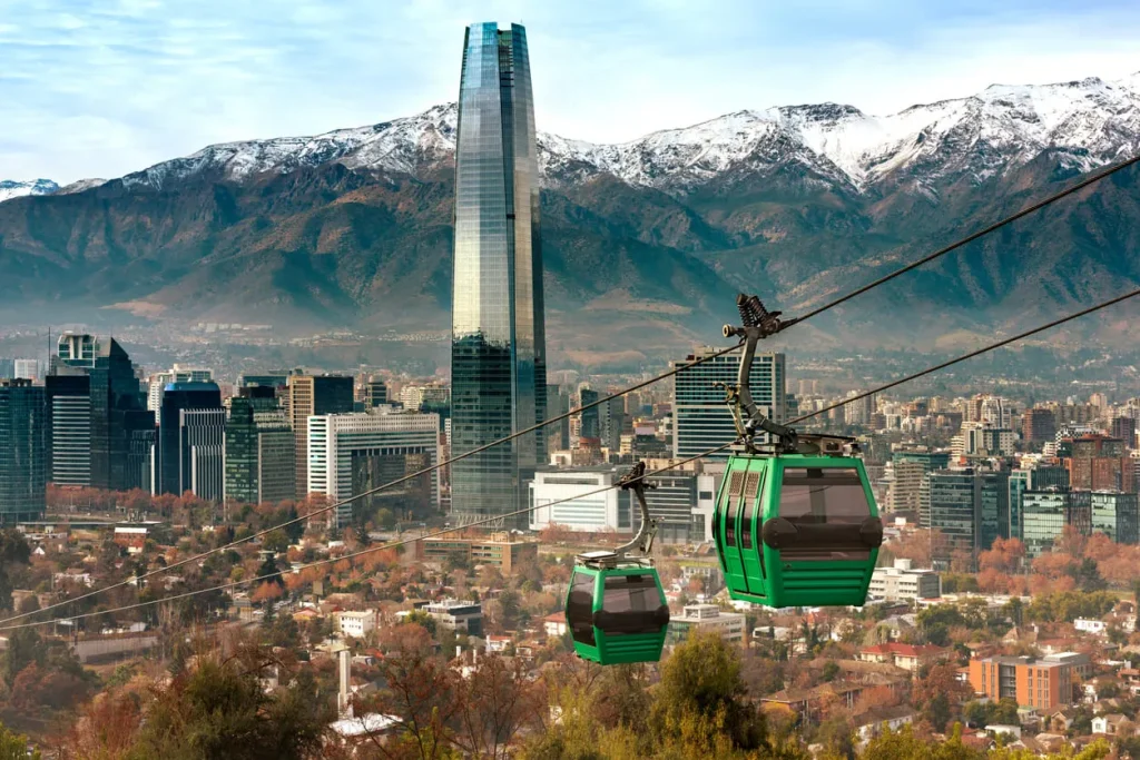 Foto dos bondinhos que fazem o circuito na cidade de Santiago, no Chile. Ao fundo, é possível ver os montes com seus picos nevados e também os prédios comerciais da região. Tem dois bondinhos em andamento.