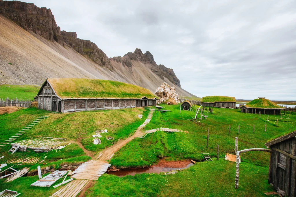 Foto de uma pequena vila viking na Escandinávia. A vegetação é bastante verde e tem uma construção extensa de madeira ao fundo. Um monte elevado fica de cenário atrás da construção.