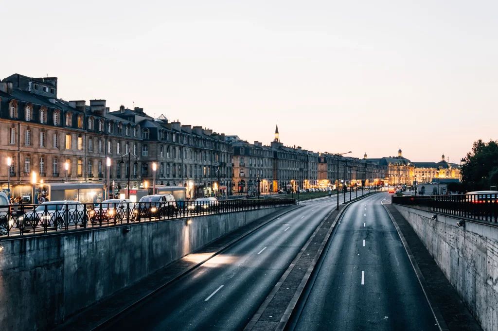 Foto do trânsito da cidade de Bordeaux, é possível ver carros parados com as luzes acesas. O céu está escurecendo e se inicia o entardecer. Tem construções nos arredores da rua bem típicas francesas.