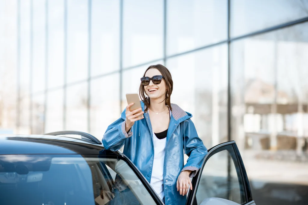 Mulher sorridente com um celular na mão. Ela está posicionada na porta no veículo, vestindo um casaco azul. O carro está parado e existe um prédio de espelhos ao fundo.