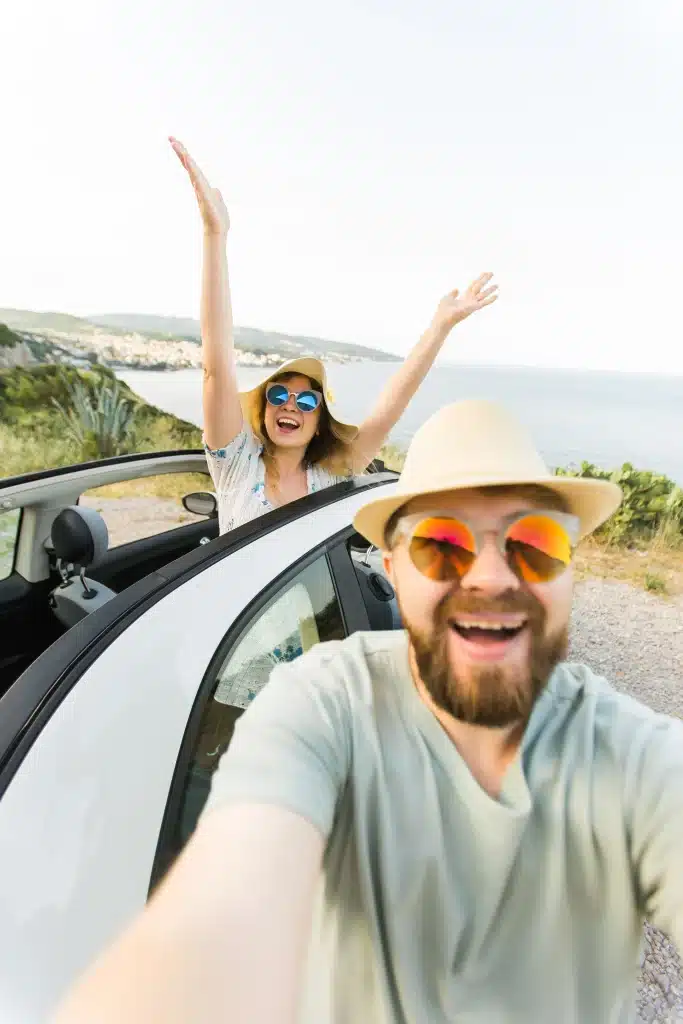 homem e e mulher felizes com o corpo para fora do carro e praia ao fundo