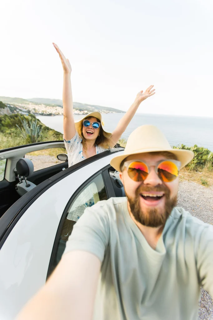 Um casal super feliz posando para uma foto selfie dentro do carro. A mulher está com os braços para o alto e sorriso no rosto. O homem está sorrindo e segurando o celular. A praia está ao fundo.