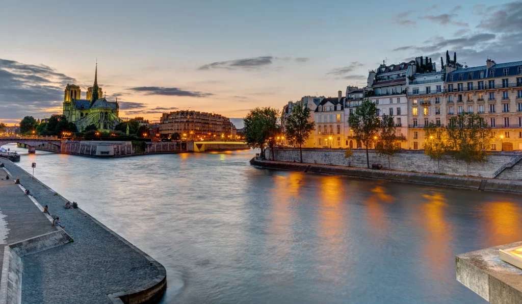 Foto noturna da cidade de Paris, na França. Ao fundo tem as casas francesas e o rio Sena passa ao meio. O céu está entardecendo.