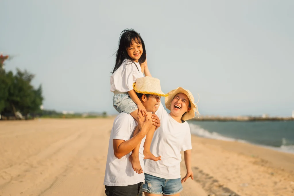 Uma família feliz na praia. A criança está sentada nos ombros do pai. A mãe brinca com a filha. A praia está ao fundo. Os três vestem camisetas brancas e shorts jeans.