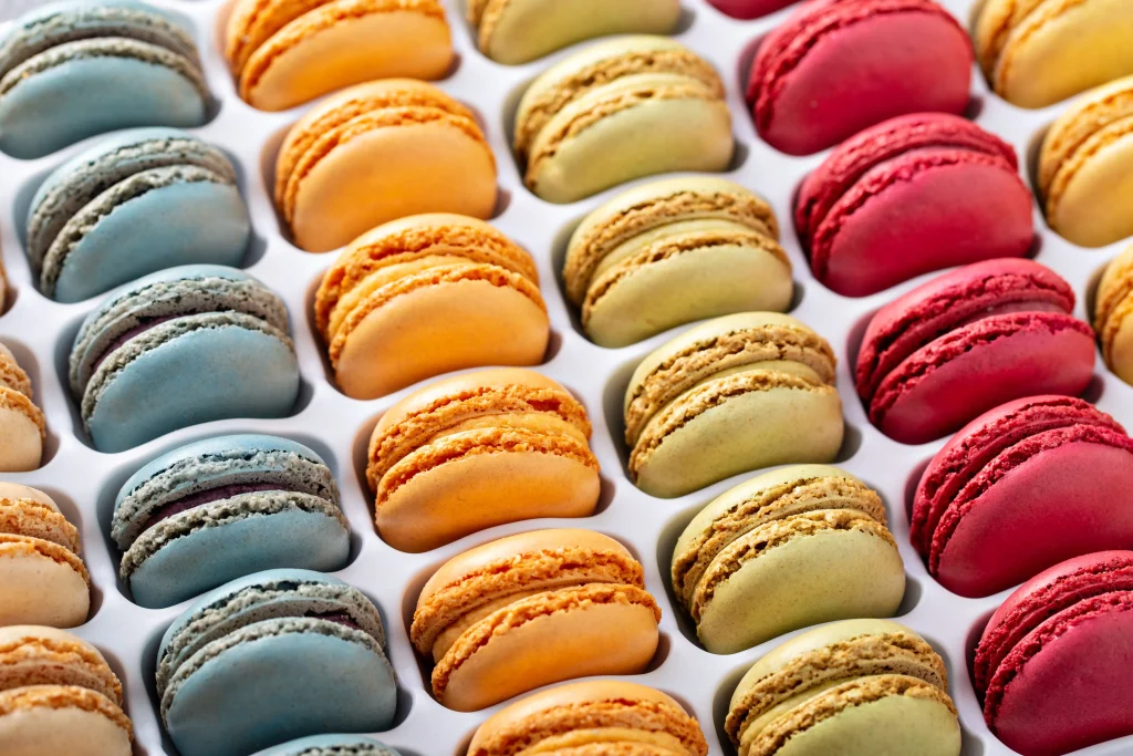 Foto de fileiras de macarons bem coloridos, doce típico da França. Parecem biscoitos recheados nas cores: vermelho, azul, laranja e amarelinho.