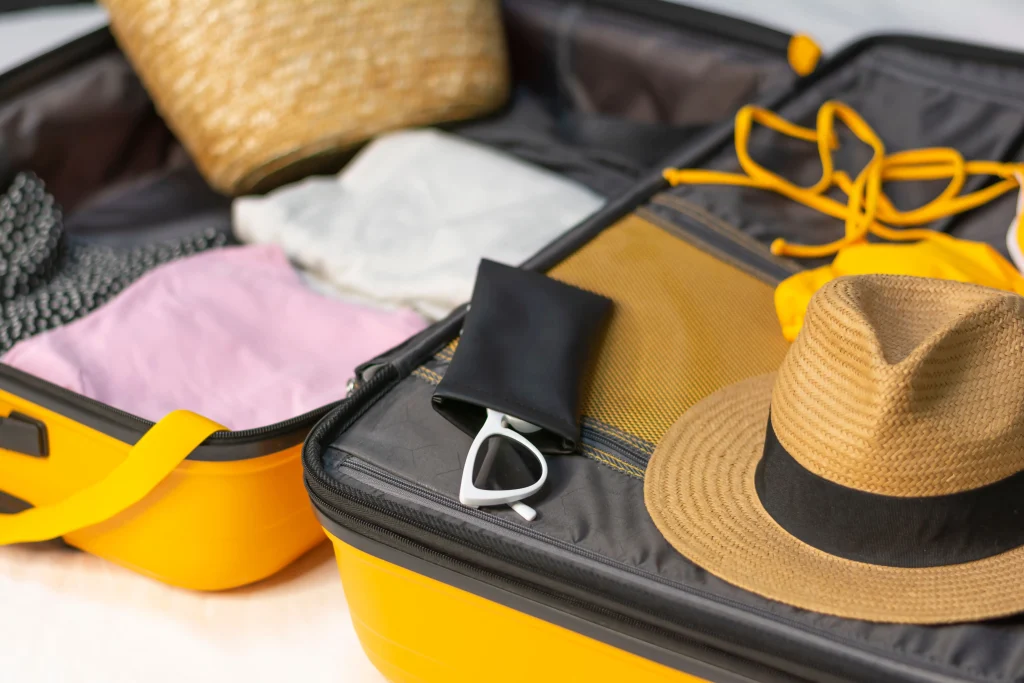 Uma mala de viagem aberta com itens de praia: chapéu, roupas, bolsa, biquini e óculos de sol.