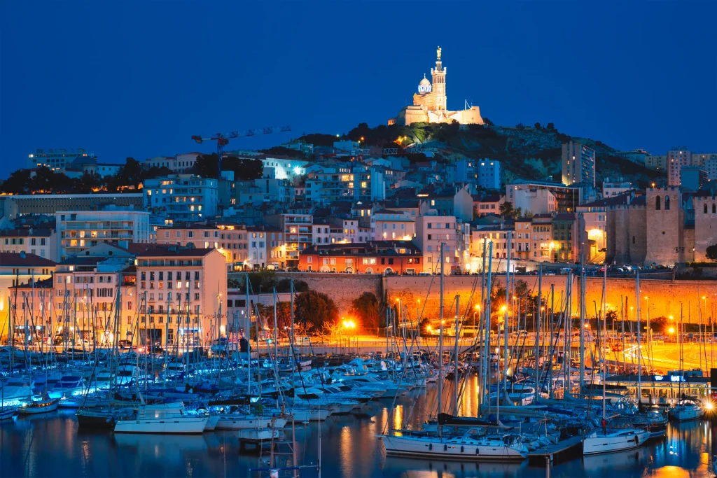 Foto noturna da cidade de Marseille, na França. No topo na montanha é possível ver um castelo bem iluminado. As casas bem típicas francesas ficam a frente, descendo o morro. Há vários barcos na marina em baixo. As luzes da cidade estão acesas.
