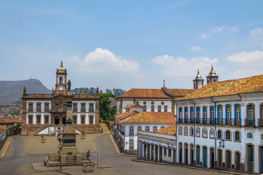 Foto de uma das igrejas mais famosas da cidade de Ouro Preto, em Minas Gerais. O solo é repleto de pedras e as casas azuis, amarelas e brancas ao redor.