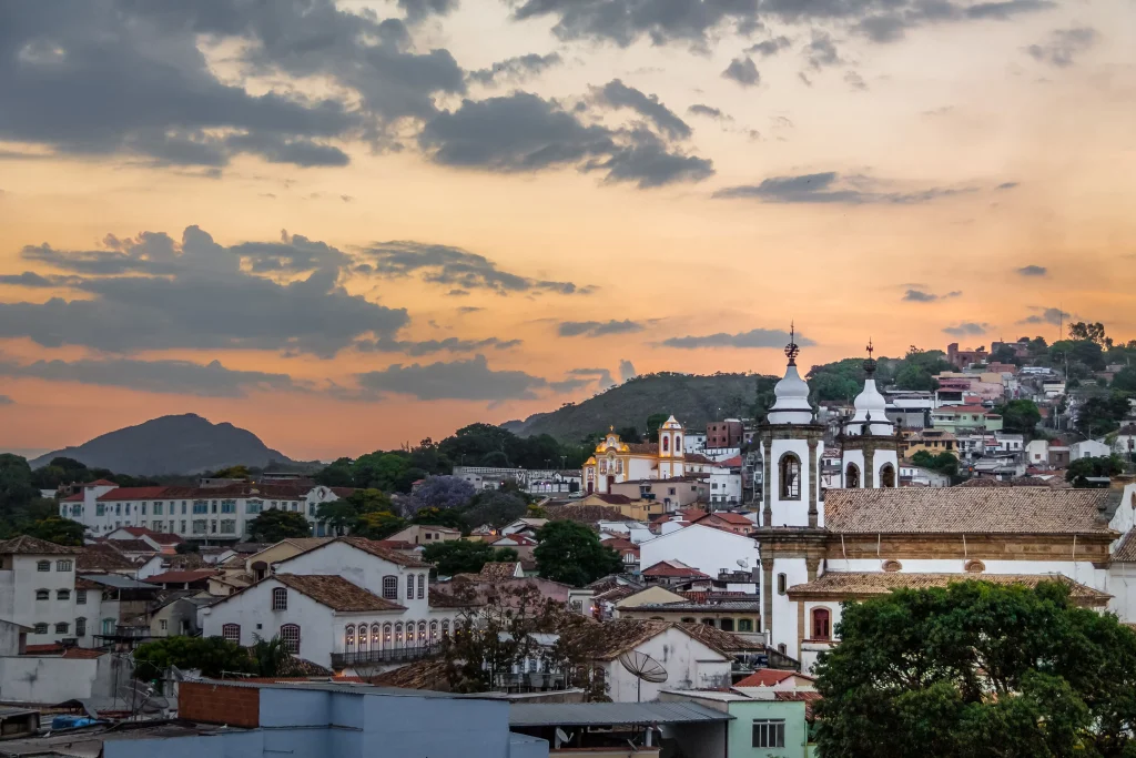 Foto aérea da cidade de São João Del Rey, em Minas Gerais. As construções são bem padronizadas na cor branca e telhas avermelhadas. O pôr do sol está acontecendo no momento da foto. É possível ver a imensidão de casas.