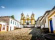 Foto de uma das igrejas mais famosas da cidade de Tiradentes, em Minas Gerais. O solo é repleto de pedras Paralelepípedos e as casas azuis, amarelas e brancas ao redor.