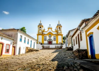 Foto de uma das igrejas mais famosas da cidade de Tiradentes, em Minas Gerais. O solo é repleto de pedras Paralelepípedos e as casas azuis, amarelas e brancas ao redor.