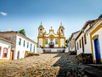 Foto de uma das igrejas mais famosas da cidade de Tiradentes, em Minas Gerais. O solo é repleto de pedras Paralelepípedos e as casas azuis, amarelas e brancas ao redor.