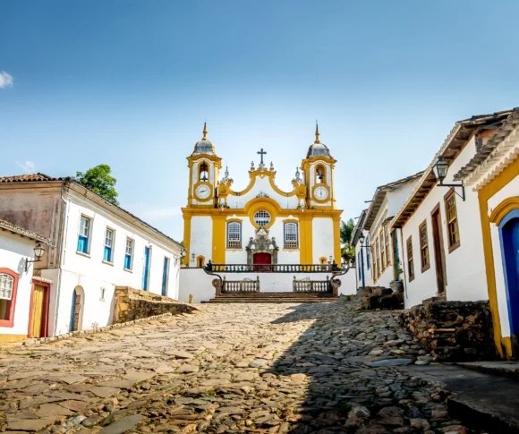 Foto de uma das igrejas mais famosas da cidade de Tiradentes, em Minas Gerais. O solo é repleto de pedras Paralelepípedos e as casas azuis, amarelas e brancas ao redor.