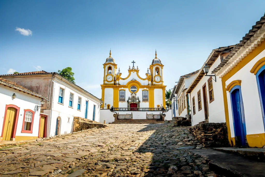 Foto de uma das igrejas mais famosas da cidade de Tiradentes, em Minas Gerais. O solo é repleto de pedras Paralelepípedos e as casas azuis, amarelas e brancas ao redor.