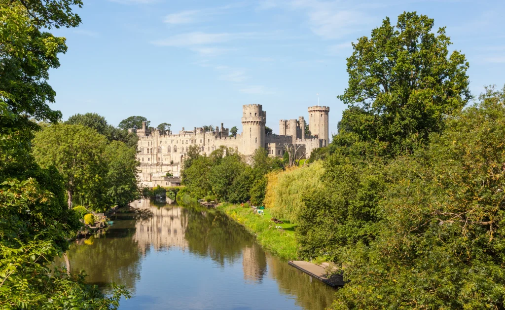 Photograph of Warwick Castle, a fortress situated on a cliff overlooking the River Avon. Green vegetation surrounds the castle grounds.
