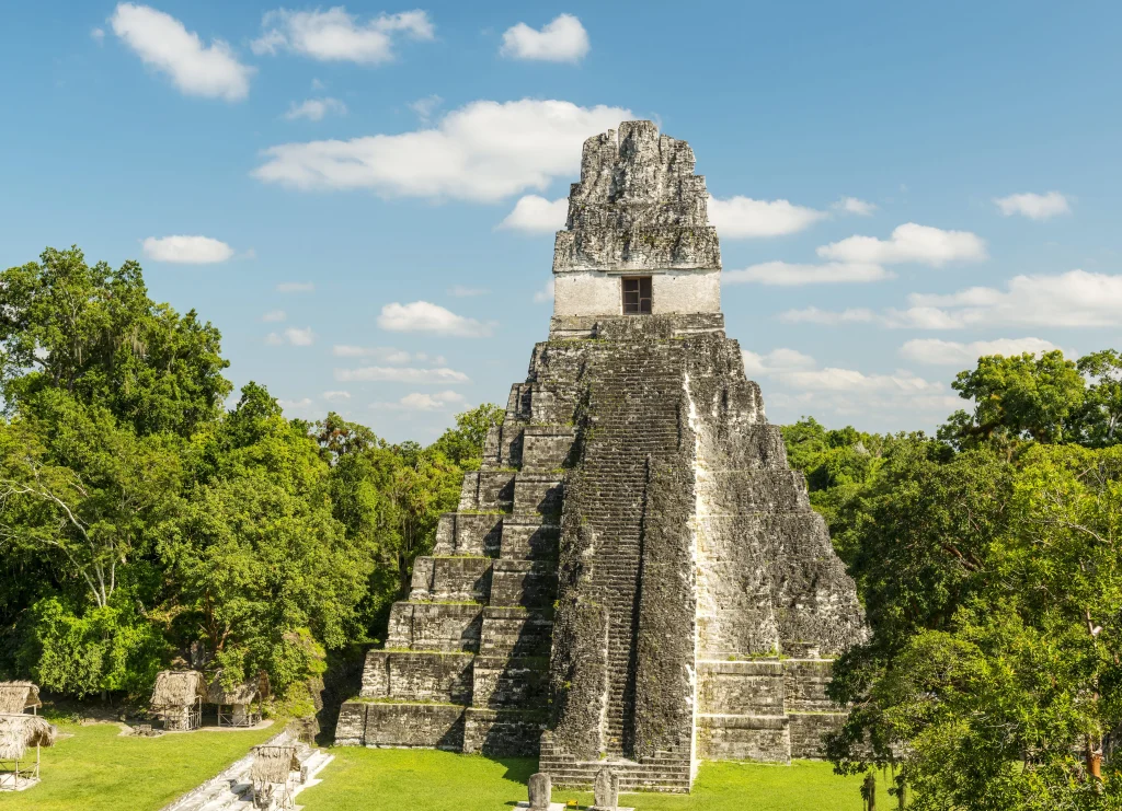 Foto do monumento Tikal na Guatemala. O céu está azul e a vegetação é bem verde. O monumento parece uma pirâmide de pedra cinza.