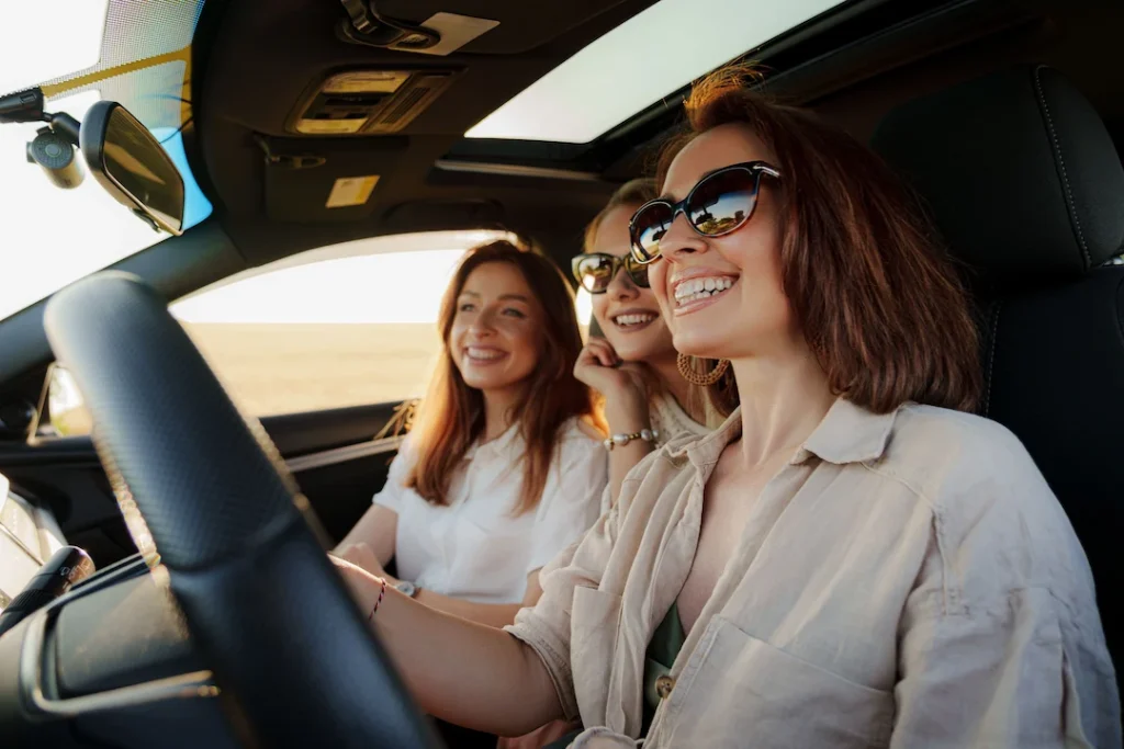 três mulheres sorrindo dentro de um carro alugado na semana do consumidor. Duas usam óculos escuros. Todas usam roupas de cores claras e tem os cabelos castanhos